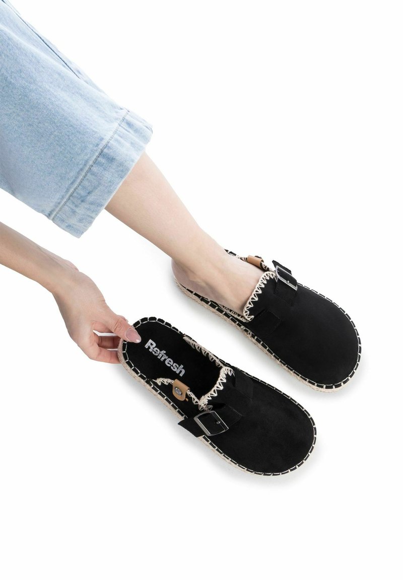 Zapatos negros de ante sin cordones con un borde cosido y una correa con hebilla. La plantilla presenta el nombre de la marca "Refresh."