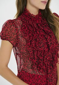 Blusa roja transparente con estampado de leopardo negro, con un escote rizado y mangas abullonadas. Botones visibles debajo de los volantes.