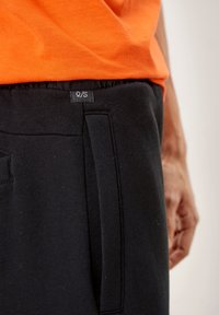 Svarta sweatpants med dragsko i midjan, med en sidficka och en etikett som visar "Q/S." Materialet verkar mjukt och texturerat.