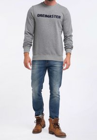 Sudadera gris con el texto "DREIMASTER", combinada con vaqueros azules y botas marrones hasta el tobillo. Presenta mangas largas y un escote redondo.