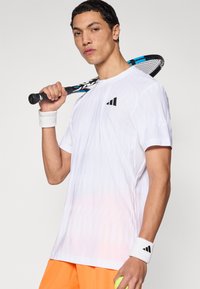 Camisa blanca de manga corta con patrón texturizado, con el logo negro de Adidas. Se lleva con pantalones cortos naranja y muñequeras blancas. Sosteniendo una raqueta de tenis.