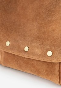 Sac en suède marron avec une fermeture à rabat ornée de trois clous en métal doré. La texture est douce avec une finition mate.