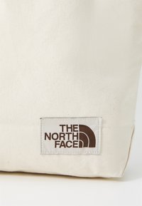 Kreminės spalvos kanvas tote krepšys su sta�čiakampiu siuvinėtu etikete, ant kurios tamsiai rudas tekstas ir logotipas. Paprastas, funkcionalus dizainas.