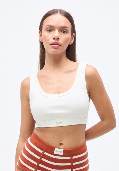 Weißes, geripptes Croptop mit weitem Ausschnitt und dünnen Trägern. Am Saum ist ein kleines Markenlabel aufgenäht. Modell kombiniert mit gestreiften Hosen.
