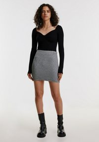 Schwarzes Langarmoberteil mit gedrehter Halslinie, kombiniert mit einem grauen karierten Minirock. Das Model trägt schwarze Stiefeletten mit einer klobigen Sohle.