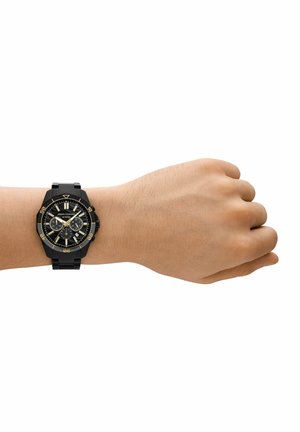 Pols met een zwart metalen Armani Exchange chronograafhorloge met gouden accenten en drie subdials op een witte achtergrond.