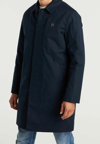 Hombre vistiendo un abrigo largo de color azul marino oscuro con un cuello y dos bolsillos frontales sobre unos jeans de color azul claro, de pie contra un fondo sencillo.