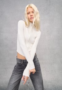 Top corto a maniche lunghe bianco a costine con colletto alto e dettaglio con zip, abbinato a jeans grigi a vita alta. Texture liscia, silhouette aderente.