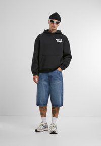 Zwarte hoodie met het "Good Days" logo, blauwe denim shorts en reflecterende sneakers. Accessoires zijn onder andere een gebreide beanie en een zonnebril.