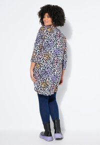 Frau mit lockigem Haar, die eine mehrfarbige Tunika mit Leopardenmuster, dunkelblaue Jeans und schwarze Stiefel mit lila Sohlen trägt, steht vor einer weißen Wand.