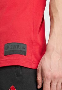 Camiseta de algodón roja con una etiqueta negra que lleva el texto "1878 MANCHESTER UNITED" y el logo de Adidas. Textura suave, manga corta.