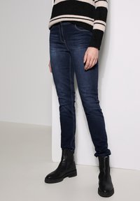 Jeans de denim ajustados en azul oscuro con un ligero estiramiento, con una cinturilla estándar y puños enrollados, combinados con botas negras hasta el tobillo.