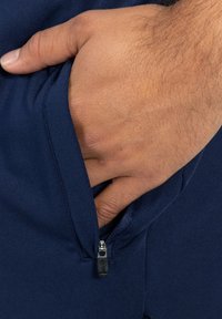 Marineblauwe ritszak op sportkleding, hand rustend binnenin, met een gladde stof textuur en een gestroomlijnd rits hardware detail.