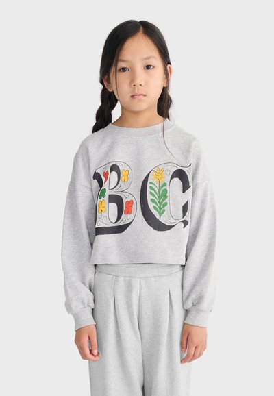 Jeune fille aux longs cheveux noirs portant un sweat-shirt gris et un pantalon, le sweat-shirt étant décoré de grandes lettres florales "B" et "C".