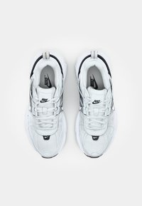 Valkoiset Nike-tennarit, joissa on teksturoitu muotoilu, mustilla yksityiskohdilla, pehmustettu nilkkaosa ja nauhat. Kengissä on paksu pohja ja logo.