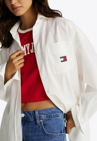 Hvid skjorte med knapper og brystlomme samt et Tommy Jeans-logo, lagdelt over en rød cropped top. Blå denimjeans synlige ved taljen.