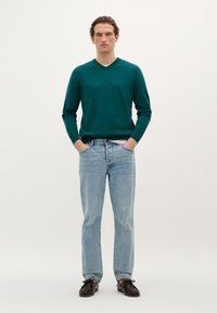 Groene v-hals trui, lange mouwen; lichtblauwe jeans; donkere schoenen. Eenvoudig ontwerp, losse pasvorm, gladde textuur zonder zichtbare patronen.