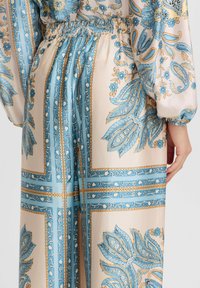 Satin-Kleid mit einem blau-goldenen Paisleymuster, elastischem Bund und Ballonärmeln, auf einem hellbeigen Stoffhintergrund.