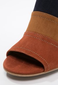 Sandale ouverte avec larges brides en daim rouille, camel et noir, avec coutures visibles et semelle beige.