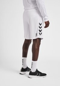 Shorts blancs avec des accents en chevrons noirs sur les côtés, fabriqués en matériau léger. Portés avec des baskets noires et des chaussettes blanches avec des rayures noires.