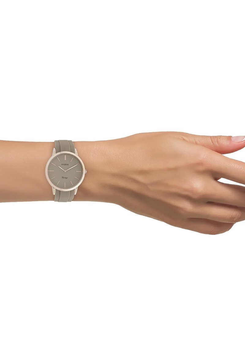 Montre ronde avec un cadran gris, design minimaliste et bracelet en silicone gris clair. Présente des index des heures fins et un boîtier en métal argenté.