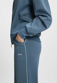 Tracksuit in soft blue fabric with zippered jacket, side pocket, and white piping. Features logo "BOSS" on the pant. Smooth texture.