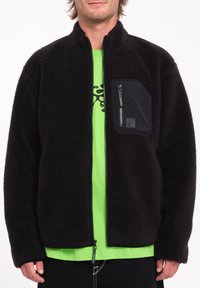 Zwarte fleece jas met een ritszak op de borst, met een gestructureerde afwerking en een groene accenten op de rits. Draagt boven een felgroen shirt.