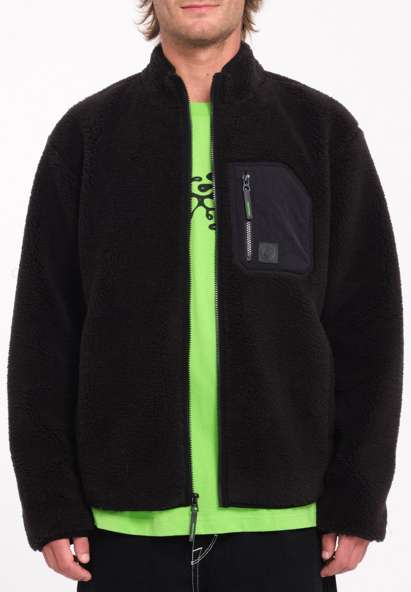 Zwarte fleece jas met een ritszak op de borst, met een gestructureerde afwerking en een groene accenten op de rits. Draagt boven een felgroen shirt.
