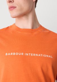 Oranžové bavlněné tričko s kulatým výstřihem a bílým nápisem "BARBOUR INTERNATIONAL" uprostřed hrudi. Hladká textura.