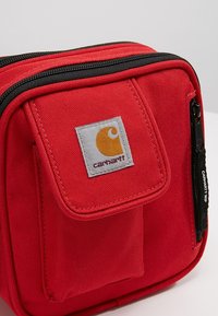 Red fabric bag with a front pocket, black zipper, and gray patch featuring a yellow logo. Textured surface and compact design.