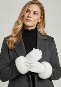 Weiße Handschuhe mit weichem, flauschigem Bündchen werden von einer Person getragen. Die Handschuhe sind glatt und eng anliegend und ergänzen einen dunklen Mantel sowie einen gerippten schwarzen Rollkragenpullover.