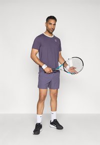 Μωβ αθλητική μπλούζα με λευκό γιακά, σε συνδυασμό με αντίστοιχα σορτς. Φορά μαύρα αθλητικά παπούτσια, λευκές κάλτσες και κρατά μια ρακέτα τένις.