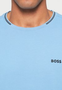 Jasnoniebieska koszulka z okrągłym dekoltem, wykonana z miękkiego materiału, z czarnymi paskami na kołnierzu i czarnym logo "BOSS" na przodzie.