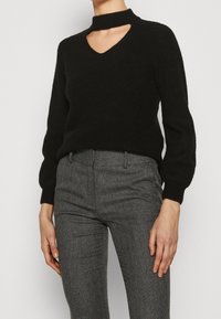 Czarny sweter z dzianiny z długim rękawem, dekoltem w szpic i wycięciem, zestawiony z wysokim stanem szarymi spodniami w jodełkę.