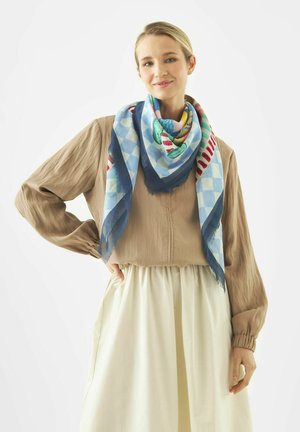 Woman with blonde hair wearing beige blouse, cream skirt, and colorful patterned scarf draped around her neck against white background.