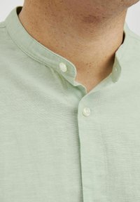 Camisa de color verde claro con cuello, confeccionada en una tela texturizada, que presenta dos botones redondos en el cuello y un diseño de corte relajado.