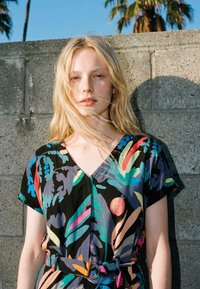 Femeie blondă cu părul liber poartă o rochie colorată cu model, stând în fața unui zid gri de beton, cu palmieri în fundal.