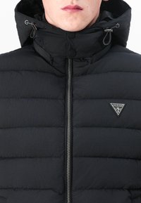 Jachetă neagră căptușită, cu guler înalt și glugă. Are fermoar pe față și un petic cu logo triangular pe piept. Textură netedă.