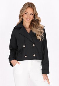 Giacca nera corta a doppio petto con ampio colletto, bottoni in argento e un leggero lucido. Abbinata a pantaloni bianchi con tasche laterali prominenti.
