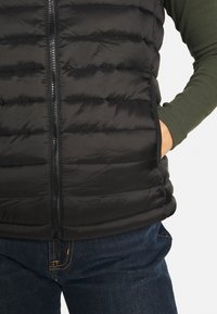 Svart puffväst med dragkedja framtill, horisontellt kviltad mönster och sidofickor, kombinerad med mörkblå jeans och en grön långärmad tröja.