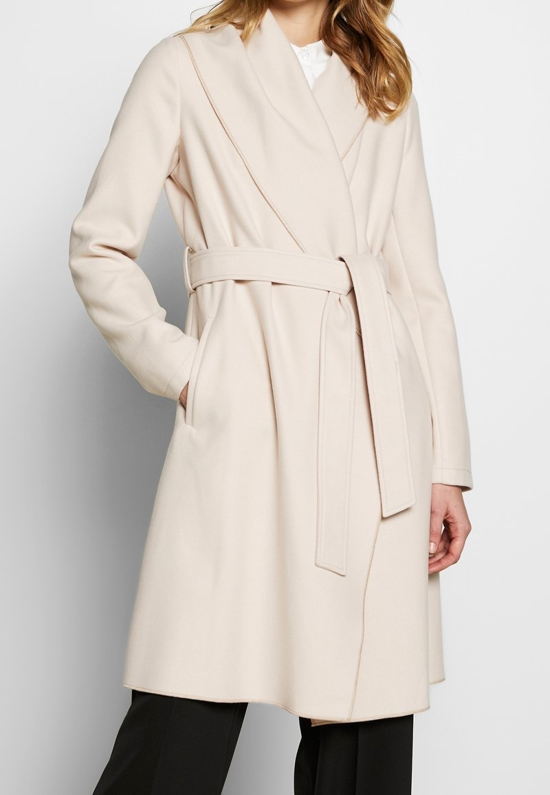 Abrigo envolvente beige hecho de una tela suave, con mangas largas, cuello profundo, bolsillos frontales y un cinturón a juego atado a la cintura.