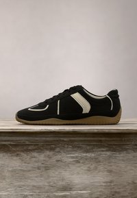Basket basse noire avec semelle beige et bandes blanches sur les côtés, exposée sur une surface en bois sur fond uni.