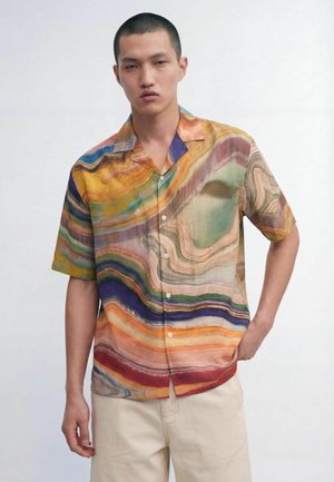 Young man with short hair wearing a short-sleeve button-up shirt with multicolored abstract wavy patterns and light beige pants.