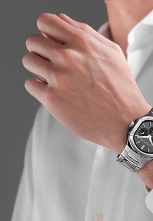 Lähikuva henkilöstä, jolla on hopeinen rannekello mustalla kellotaululla ja metallirannekkeella, pukeutunut valkoiseen paitaan.