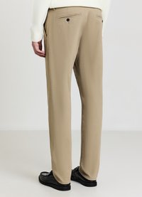 Pantaloni beige con una texture liscia, vestibilità sartoriale e una tasca posteriore con un solo bottone. Indossati con scarpe nere.