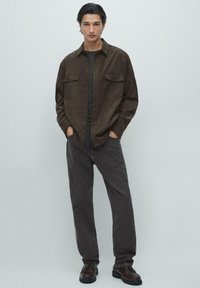 Młody mężczyzna ubrany w brązową koszulę z guzikami na ciemnym t-shircie, ciemnoszare dżinsy i brązowe buty sznurowane, stojący z rękami w kieszeniach.