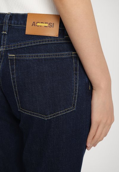 Mørkeblå denimjeans med gul søm, med et lærmerke merket "ACRESI" på livet og en design på baklommen.