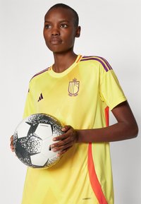 Camisola atlética amarelo-brilhante com acentos roxos, apresentando um padrão diagonal e um emblema. O jogador segura uma bola de futebol em preto e branco.