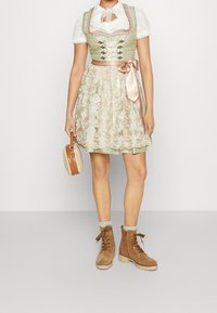 Robe traditionnelle verte et crème avec détails en dentelle, ruban en satin et manches bouffantes courtes, associée à des bottines marron à lacets et un sac à main rond.