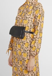 Borsa a tracolla in pelle nera con finitura liscia, forma rettangolare e chiusura a pattina, indossata sopra un vestito con motivo floreale e accenti gialli.